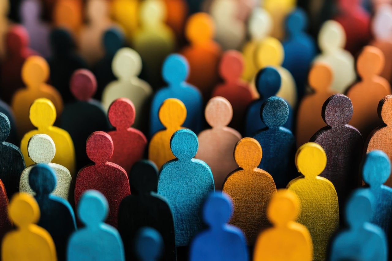 Close-up image of a large, diverse crowd of stylised wooden human figures painted in various colours, representing diversity and inclusion. The figures are grouped closely together, symbolising community, public participation, and collective voice.