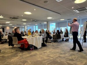 Professor Christopher Kipps, the Wessex Secure Data Environment lead, stands beside a flipchart speaking to a room of participants seated at round tables during the final day of the Wessex Public Panel on NHS Data. The group listens attentively, with posters and discussion materials displayed around the bright, modern meeting room.