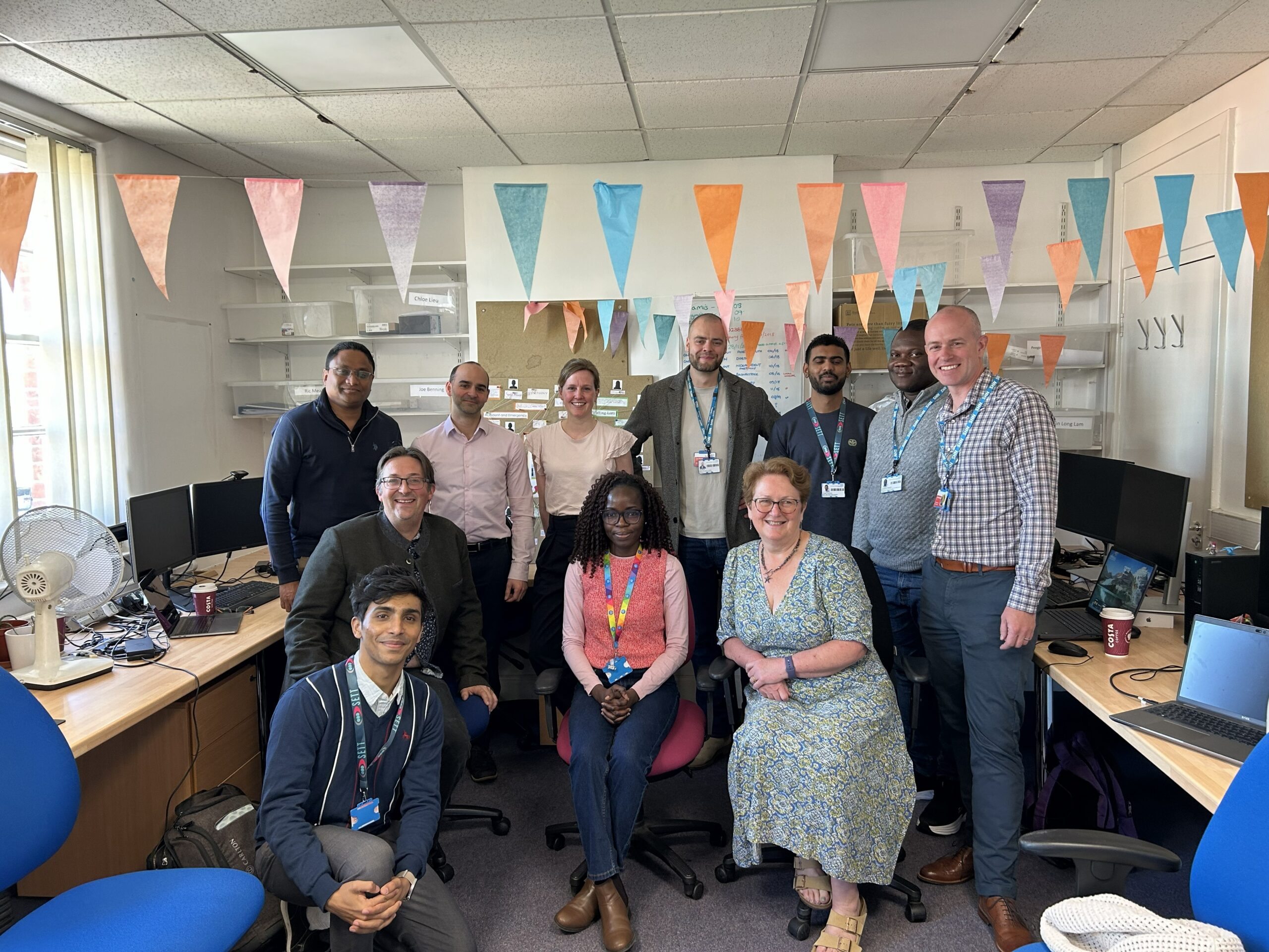 The GAP-S study Hack Day team gathered for a group photograph during a collaborative workshop. Eleven team members are pictured together in an open-plan office decorated with colourful bunting.